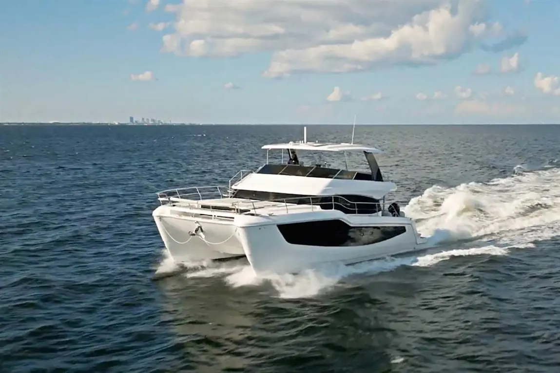 A luxury catamaran sailing on calm waters, leaving a wake, with a skyline visible in the distance under a blue sky.